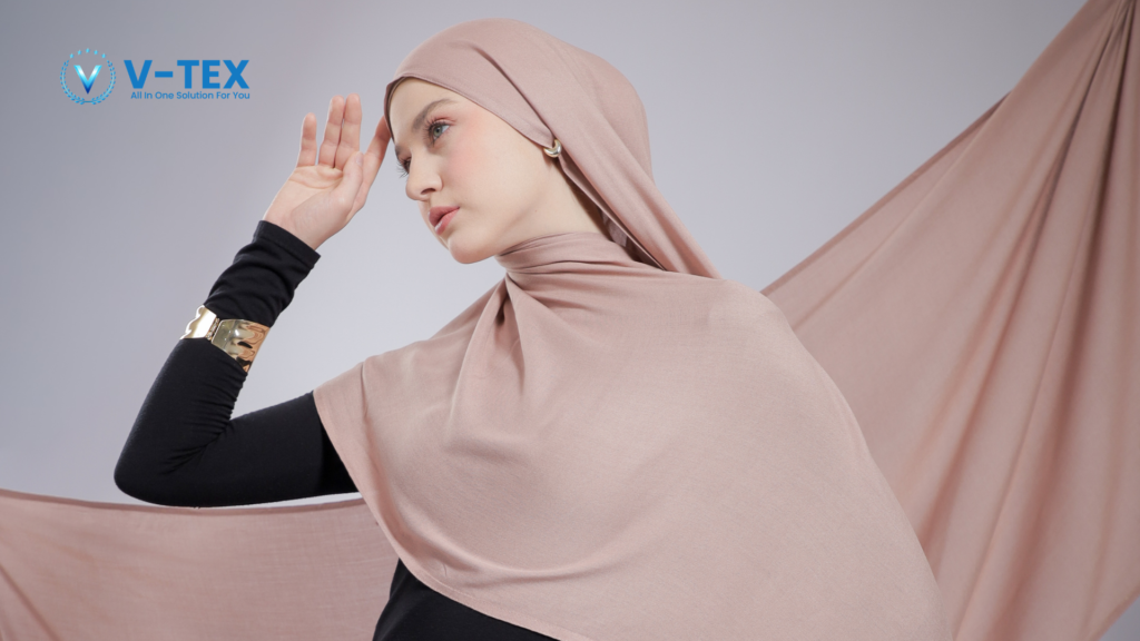 Model wanita berpose dari samping mengenakan hijab polos berwarna dusty pink berbahan kain viscose yang tampak lembut dan jatuh. Model memadukan hijab tersebut dengan busana dalaman (manset) hitam serta aksesori gelang emas, berlatar belakang studio dengan dekorasi bentangan kain viscose senada. Di sudut kiri atas terdapat logo V-TEX.